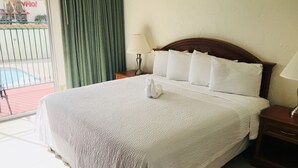 A hotel room with a large bed, a wooden headboard, a nightstand with a lamp, and a view of a pool and building outside.