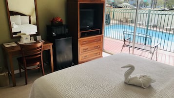 A hotel room with a bed, a wooden dresser, a television, a desk with a lamp, and a view of a swimming pool.