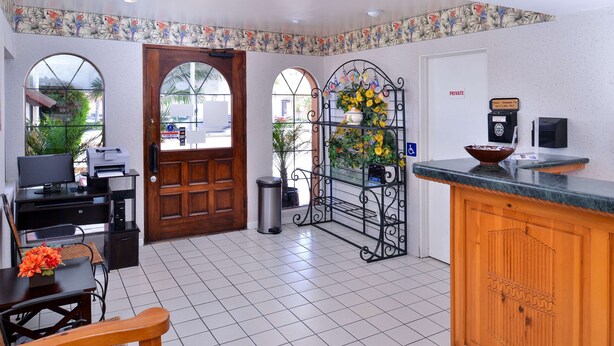A reception area with a wooden desk, a computer, and a coffee machine. There is a door with a 'PRIVATE' sign, a trash can, and a decorative metal rack with flowers. - Americas Best Value Inn Loma Lodge (San Diego)