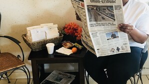 A person seated at a table reading a newspaper, with a basket of items, a cup, and a flower on the table. - Americas Best Value Inn Loma Lodge (San Diego)