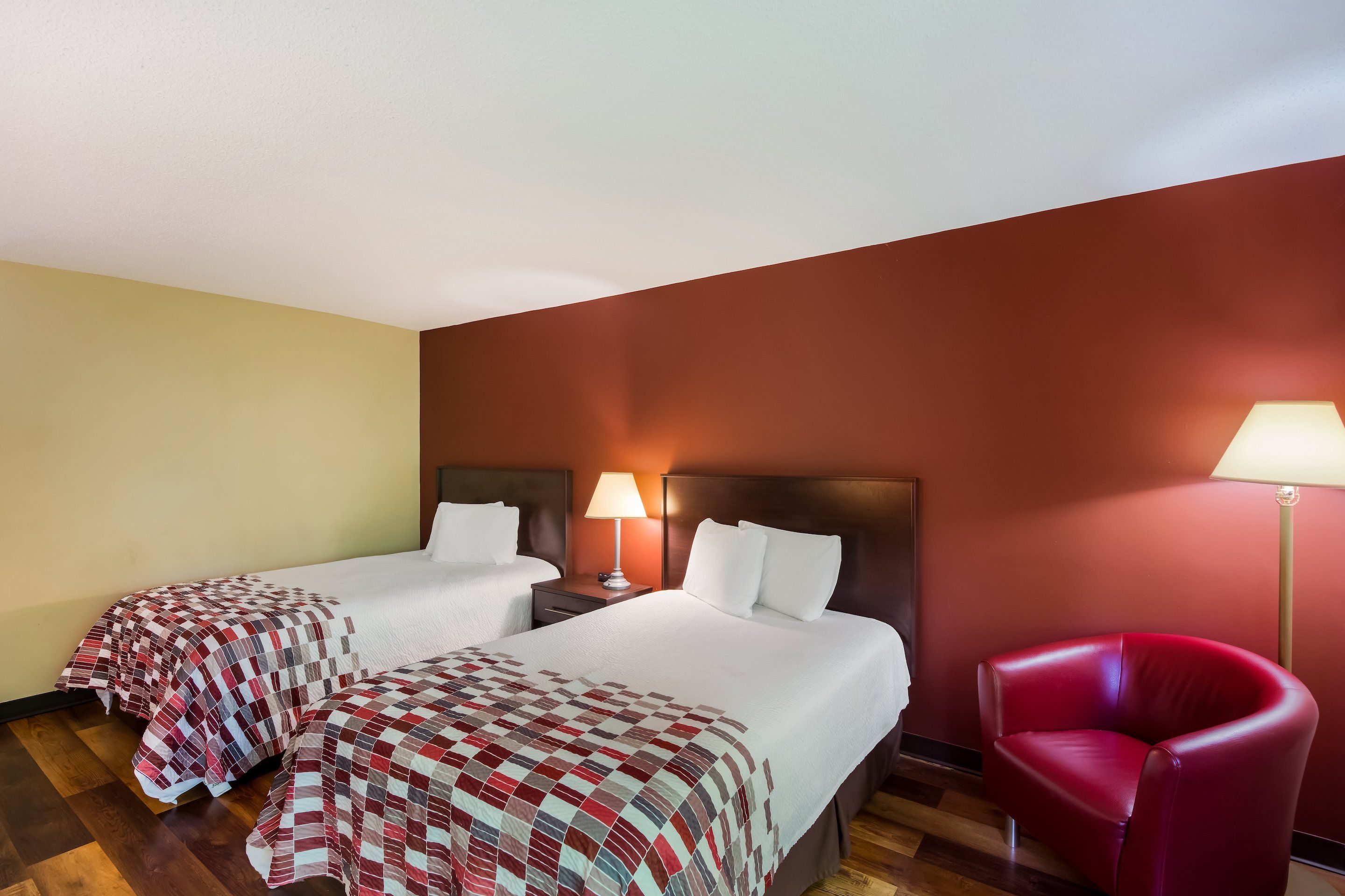 Una habitación de hotel con dos camas, una silla roja y una mesita de noche con lámpara.