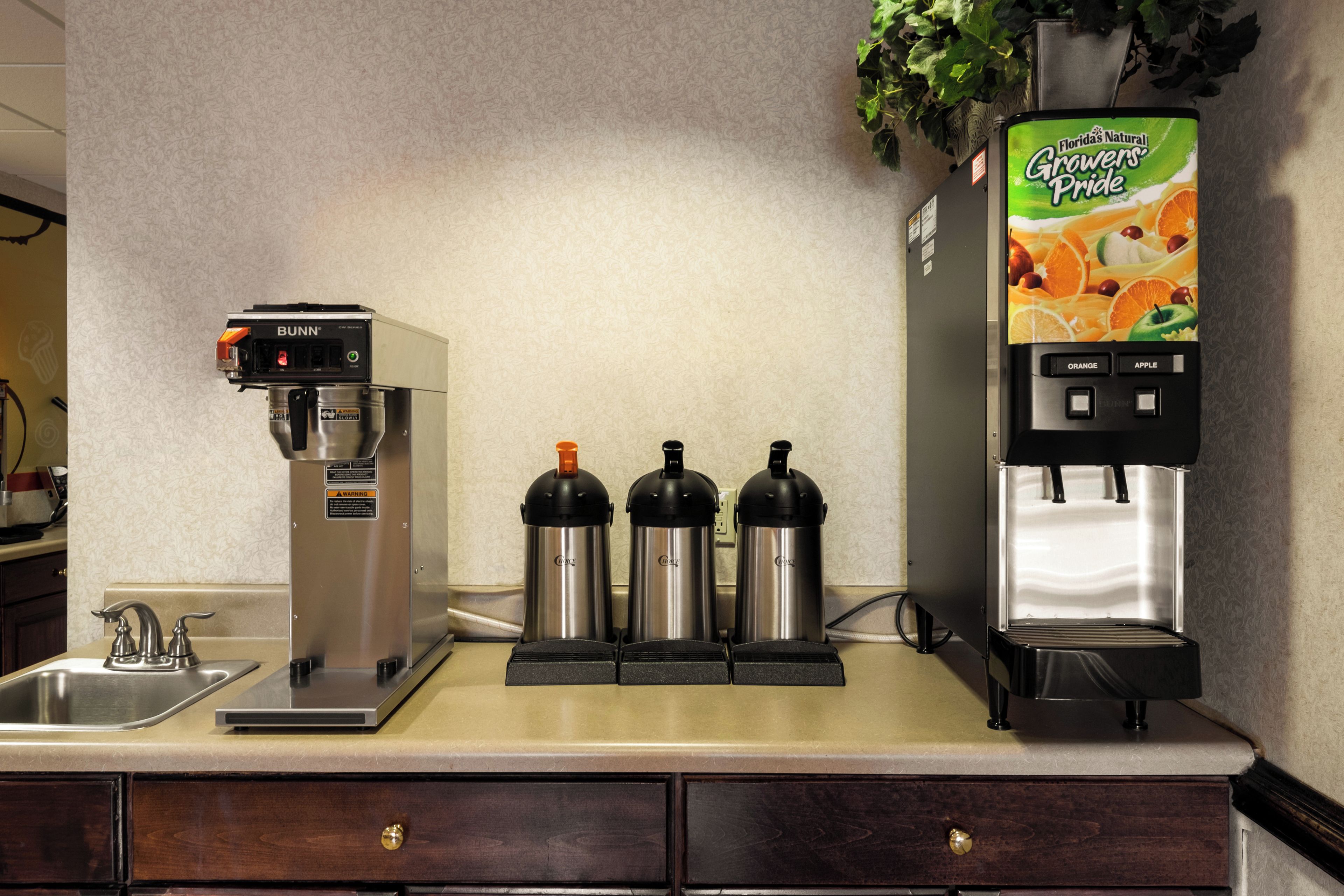 Una estación de café con una máquina de café Bunn, tres tazas térmicas y un dispensador de jugo.