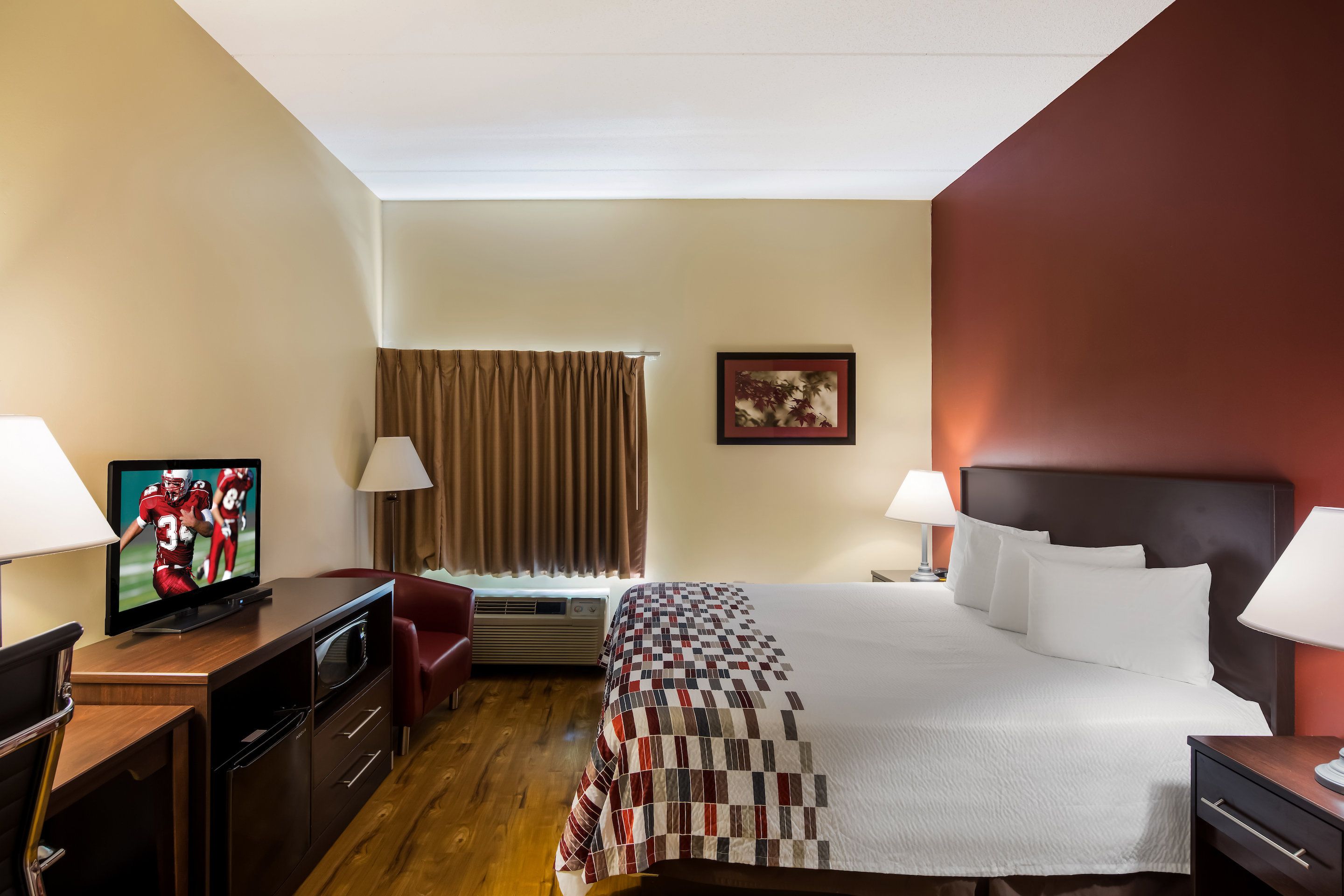 Habitación de hotel con cama, dos lámparas de noche, televisor sobre un escritorio, una silla, ventana con cortinas e imagen enmarcadas en la pared.