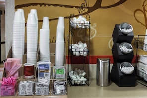 On voit une variété de gobelets jetables, des sachets de sucre et un thermos posés sur un comptoir.