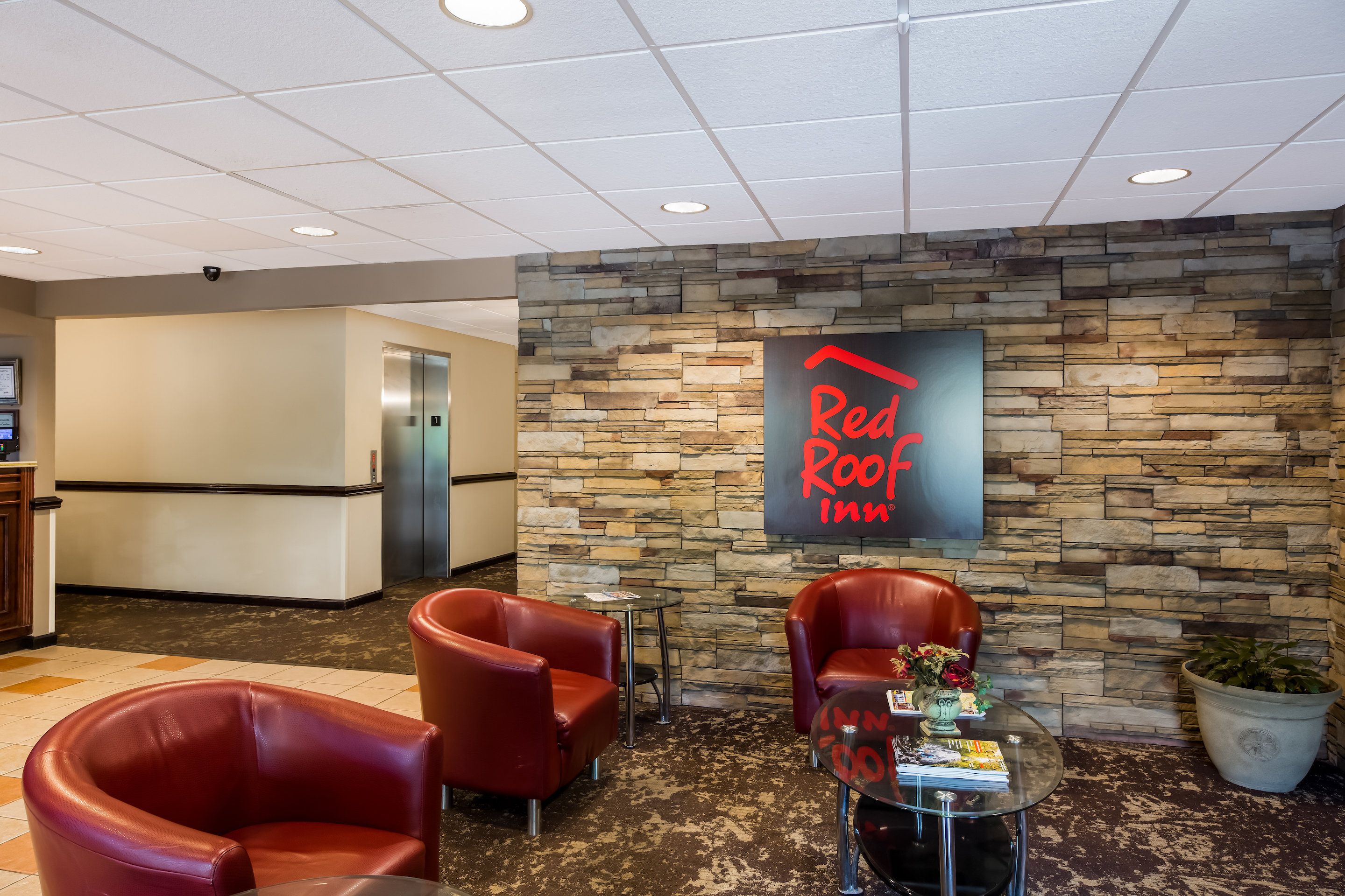 A Red Roof Inn lobby with stone walls, leather chairs, a glass table with magazines, and an elevator.
