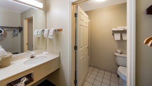 Une salle de bain avec un lavabo, un miroir et des toilettes. On y trouve un porte-serviettes avec des serviettes et une porte de douche.