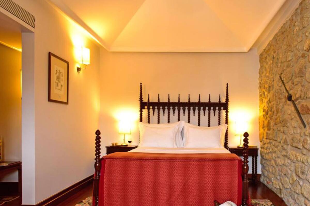 A hotel room with a bed, two bedside lamps, a stone wall, and a framed picture on the wall.