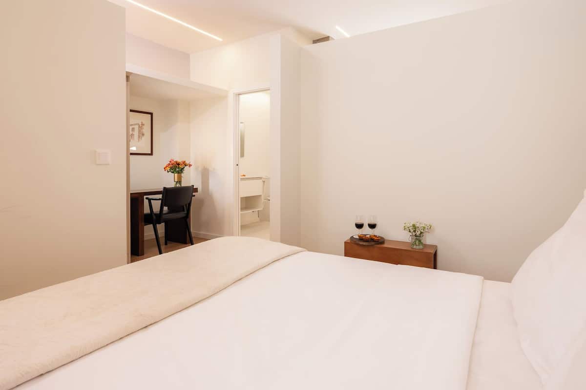 A hotel room with a bed, a desk with a chair, a small table with two glasses of wine, and a vase with flowers.