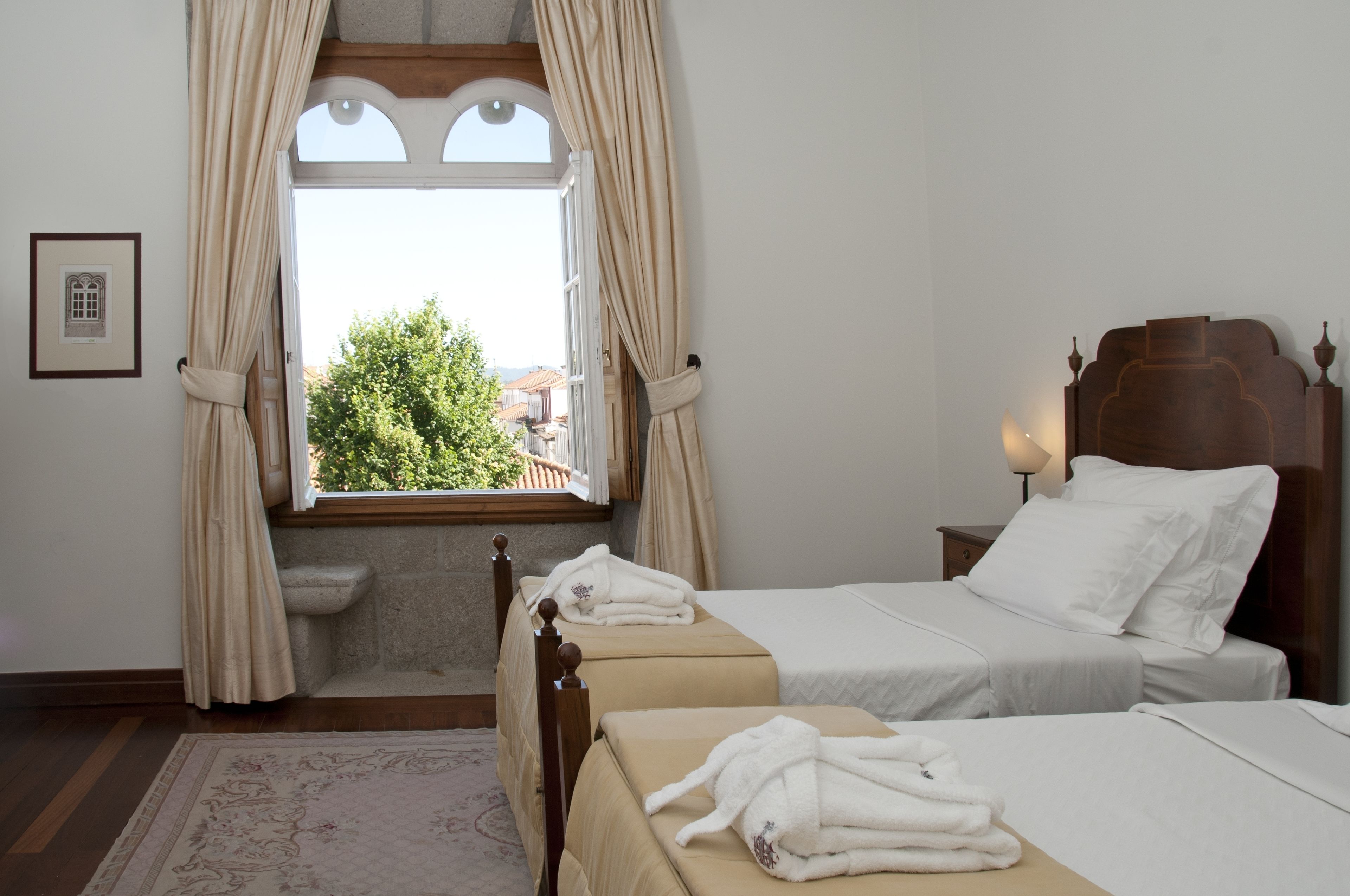 A hotel room with two beds, a window with curtains, a wooden headboard, a nightstand with a lamp, and a framed picture on the wall.