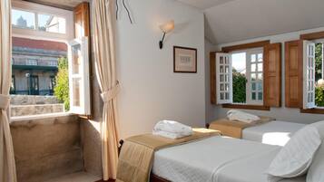 Two single beds with white linens and a throw blanket, a nightstand with a lamp and framed picture, and a window with a view.