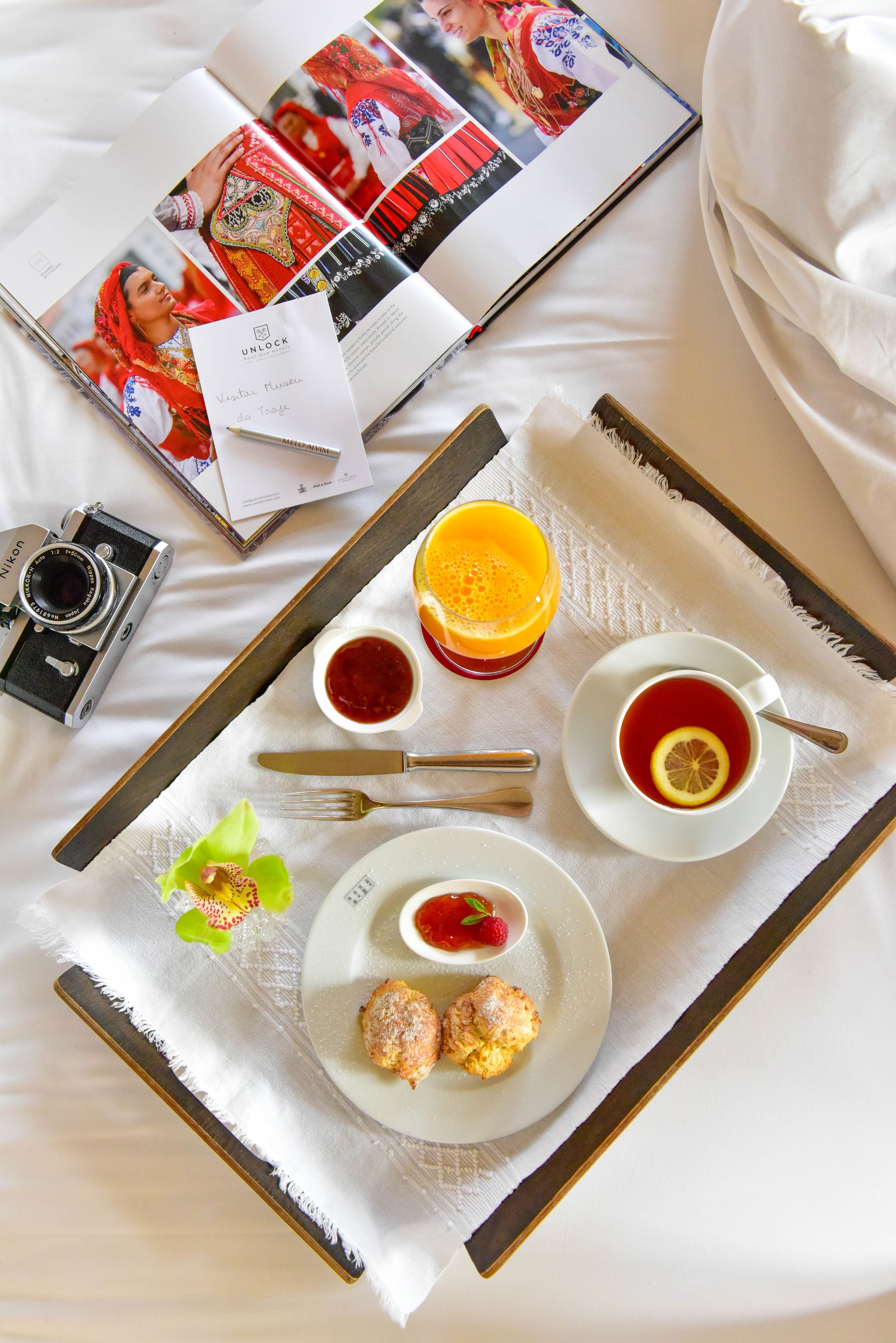 A neatly arranged breakfast tray with a camera, a magazine, and a book on a bed.