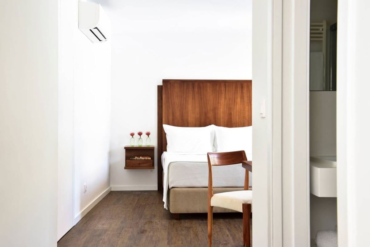 A bedroom with a wooden headboard, a bed with white linens, a bedside table with a vase, and a chair.