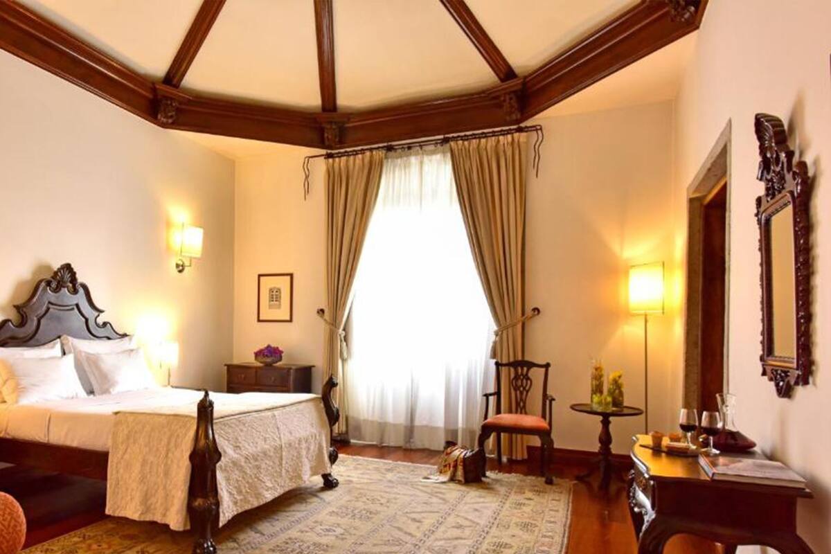 A hotel room with a large bed, two bedside tables, a chair, a small table with a lamp, a mirror, and a window with curtains.