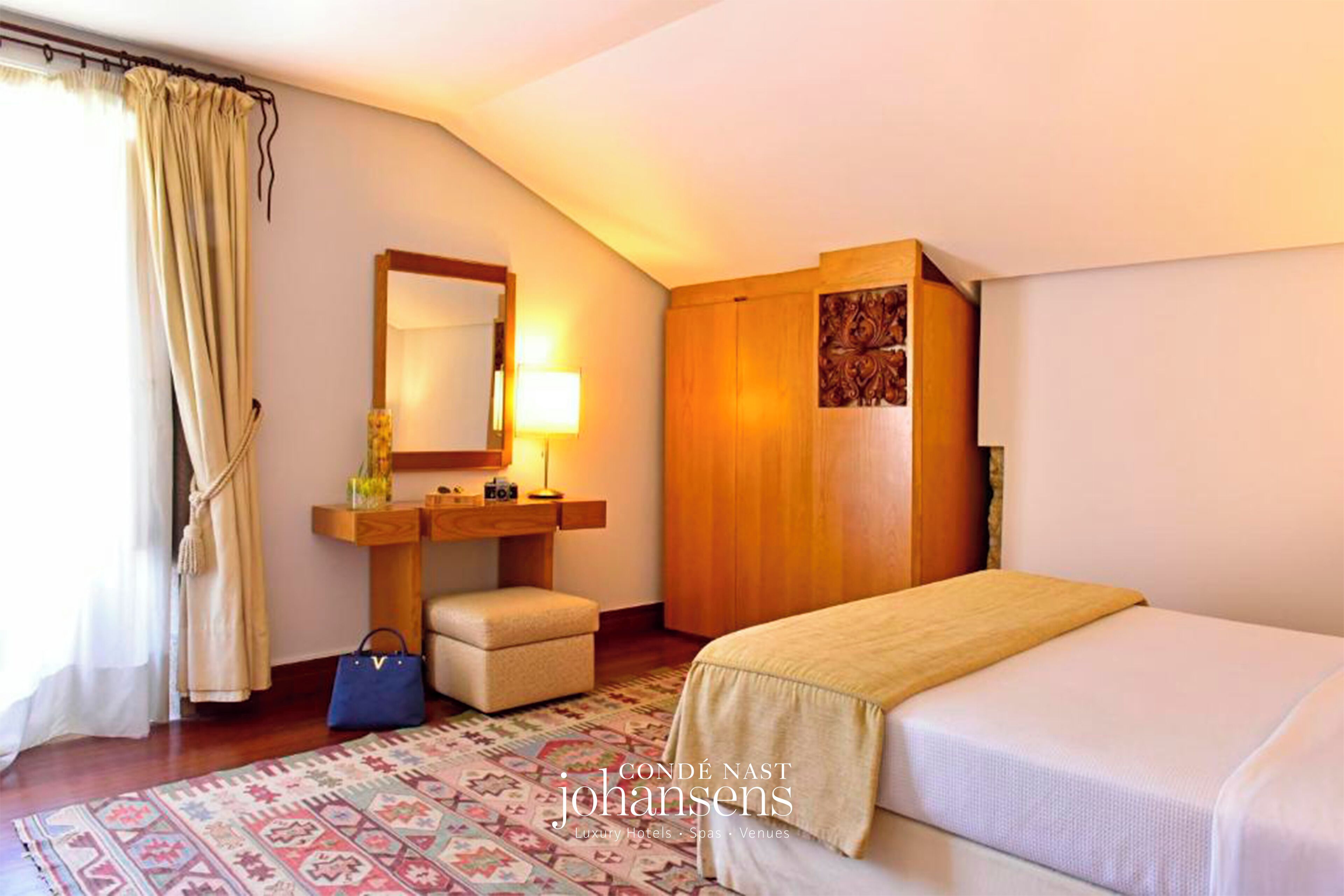 Een hotelkamer met een bed, nachtkastje, lamp, spiegel, houten garderobe en een kamerbreed tapijt met dessin.