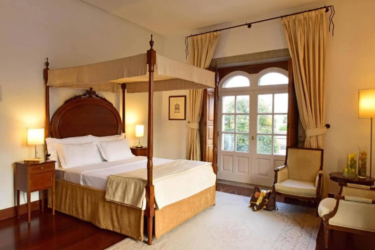 A four-poster bed with a canopy, a sitting chair, a small table with a lamp, and a view of the outdoors through a window.