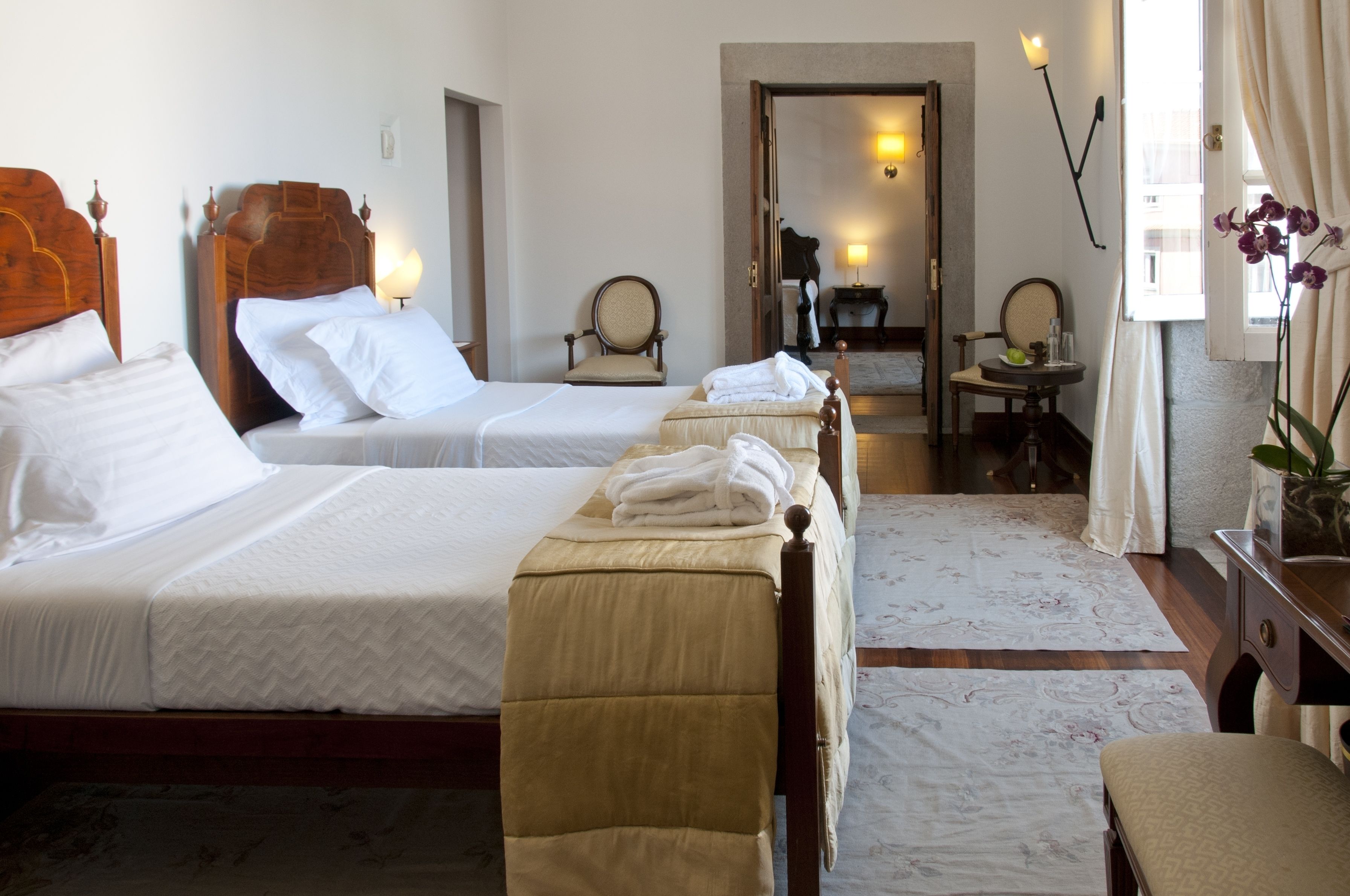 A hotel room with two single beds, a sitting area with a chair and table, and a view of the street through an open door.
