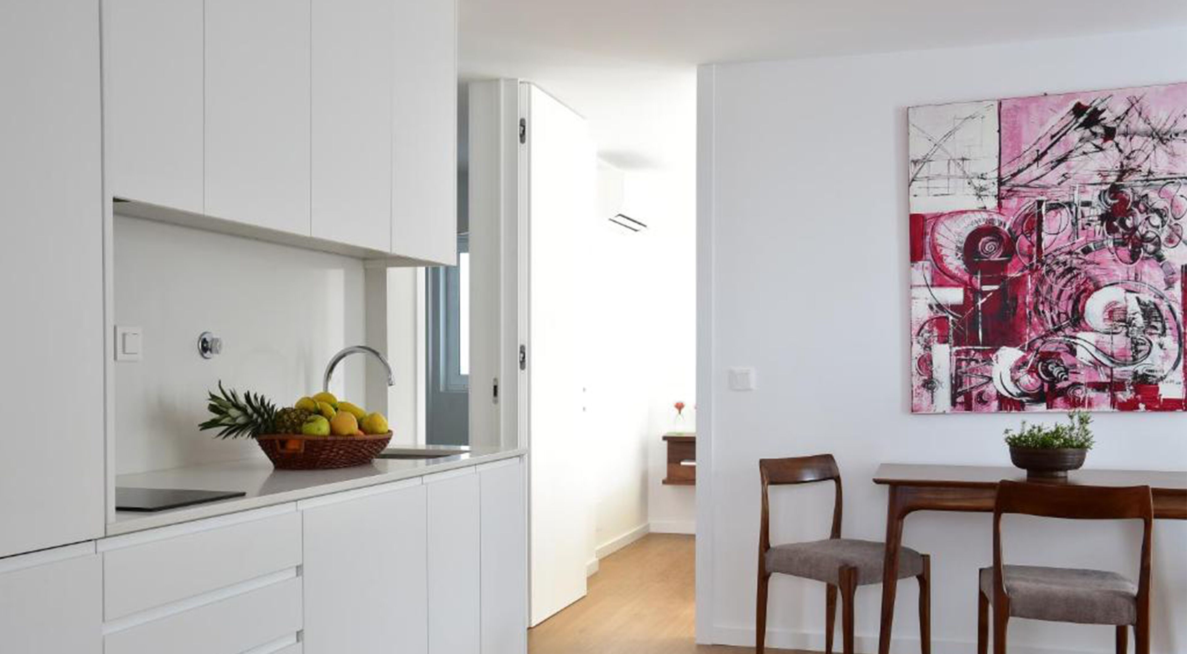 Una cocina moderna con armarios blancos, una cesta de frutas y un comedor con dos sillas y una mesa.