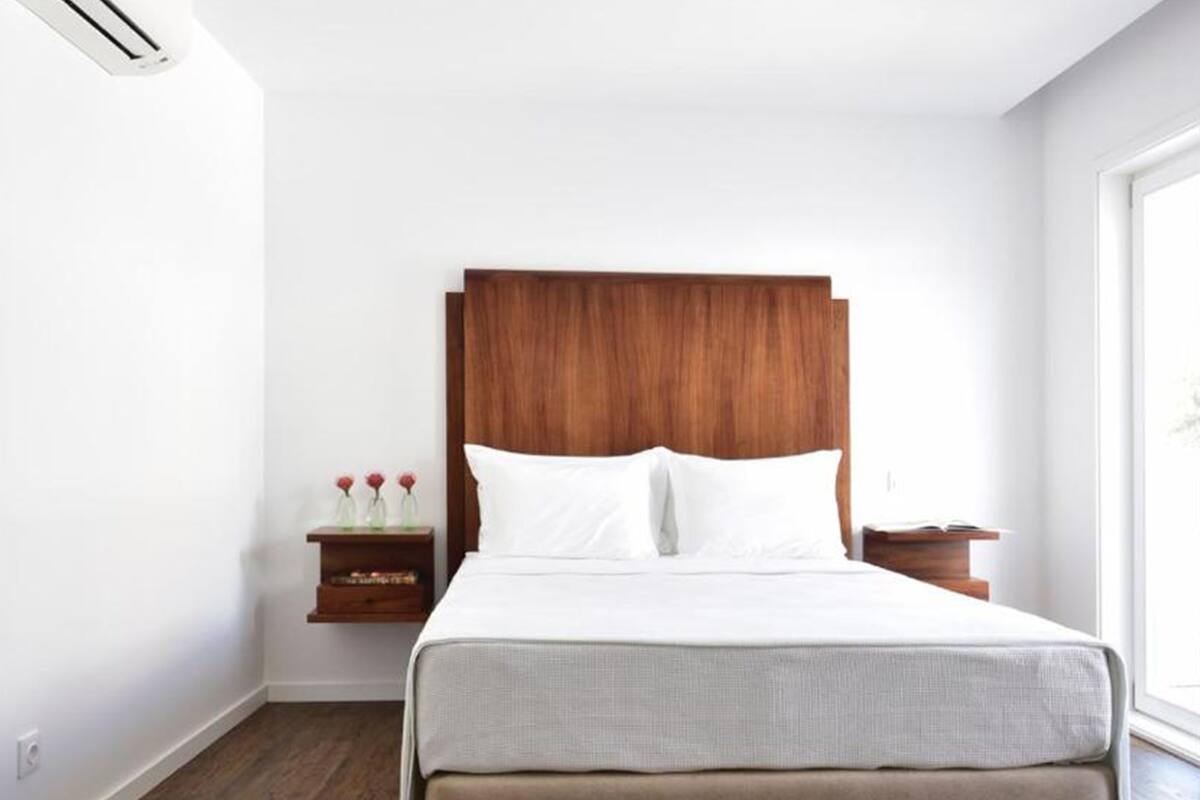 A hotel room with a large bed, wooden headboard, white bedding, a nightstand with a vase of flowers, and a window providing natural light.