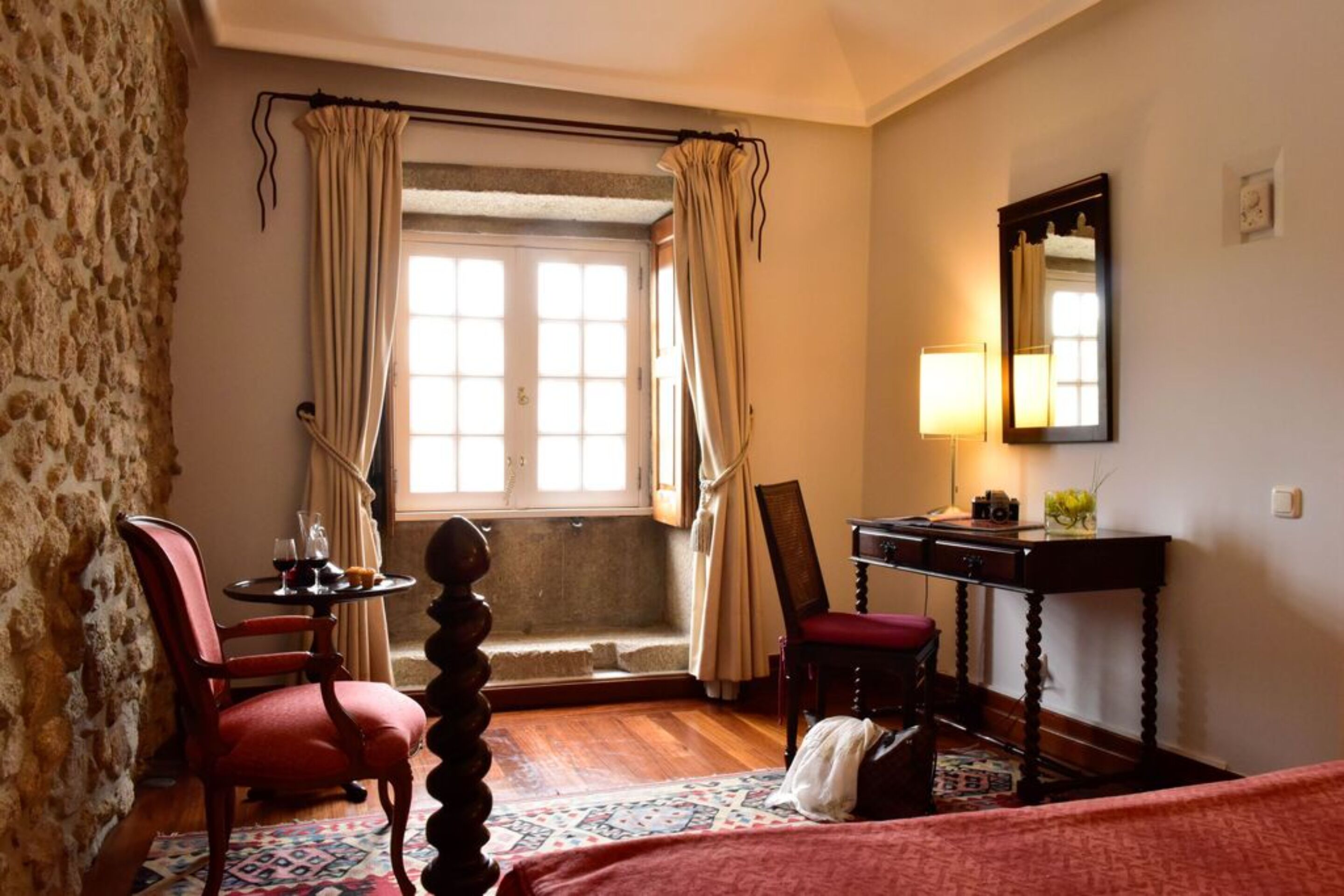 Habitación de hotel con pared de piedra, cama con colcha roja, escritorio de madera con lámpara, una silla y una ventana con cortinas.
