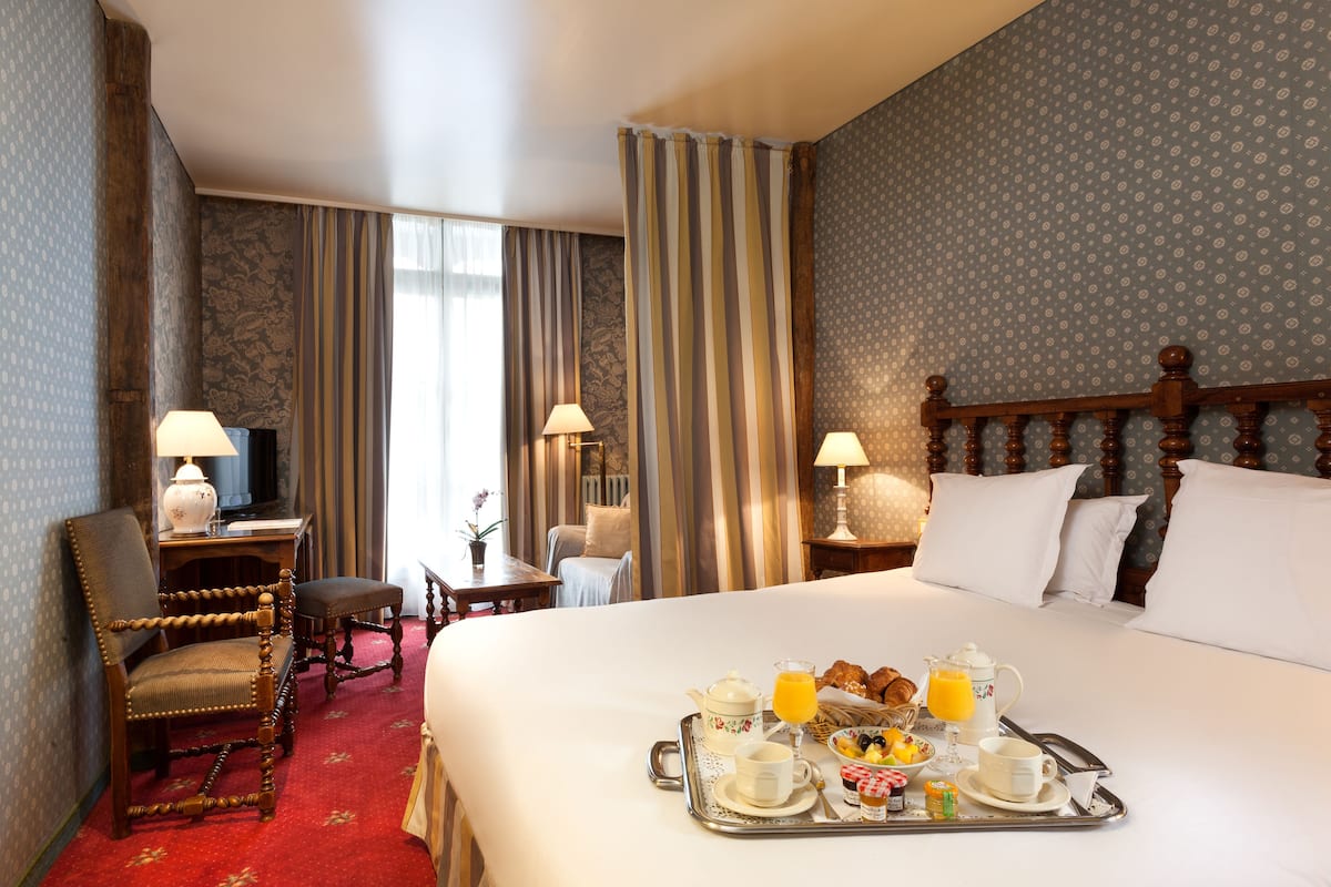 A hotel room with a large bed, a wooden chair, a desk with a lamp, and a breakfast tray on the bed.