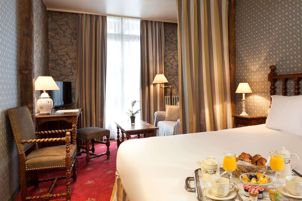 A hotel room with a large bed, a desk with a lamp, a chair, a small table with a vase, and a breakfast tray with coffee, juice, and pastries.