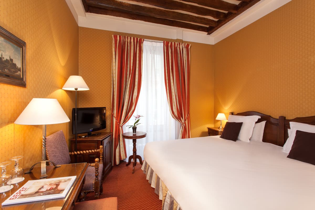 A hotel room with a bed, a television, a desk with a lamp, a chair, and a small table with a flower vase.