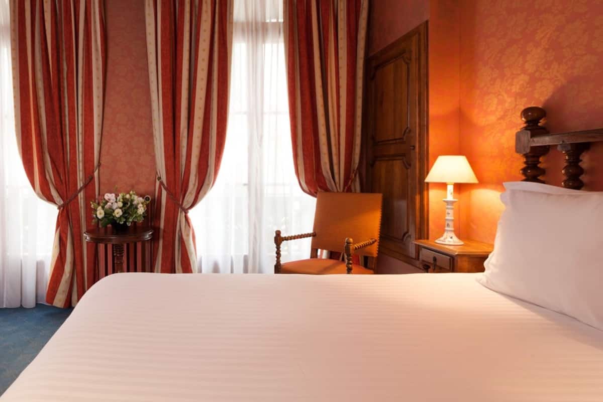 A hotel room with a large bed, a wooden chair, a bedside table with a lamp, and curtains.
