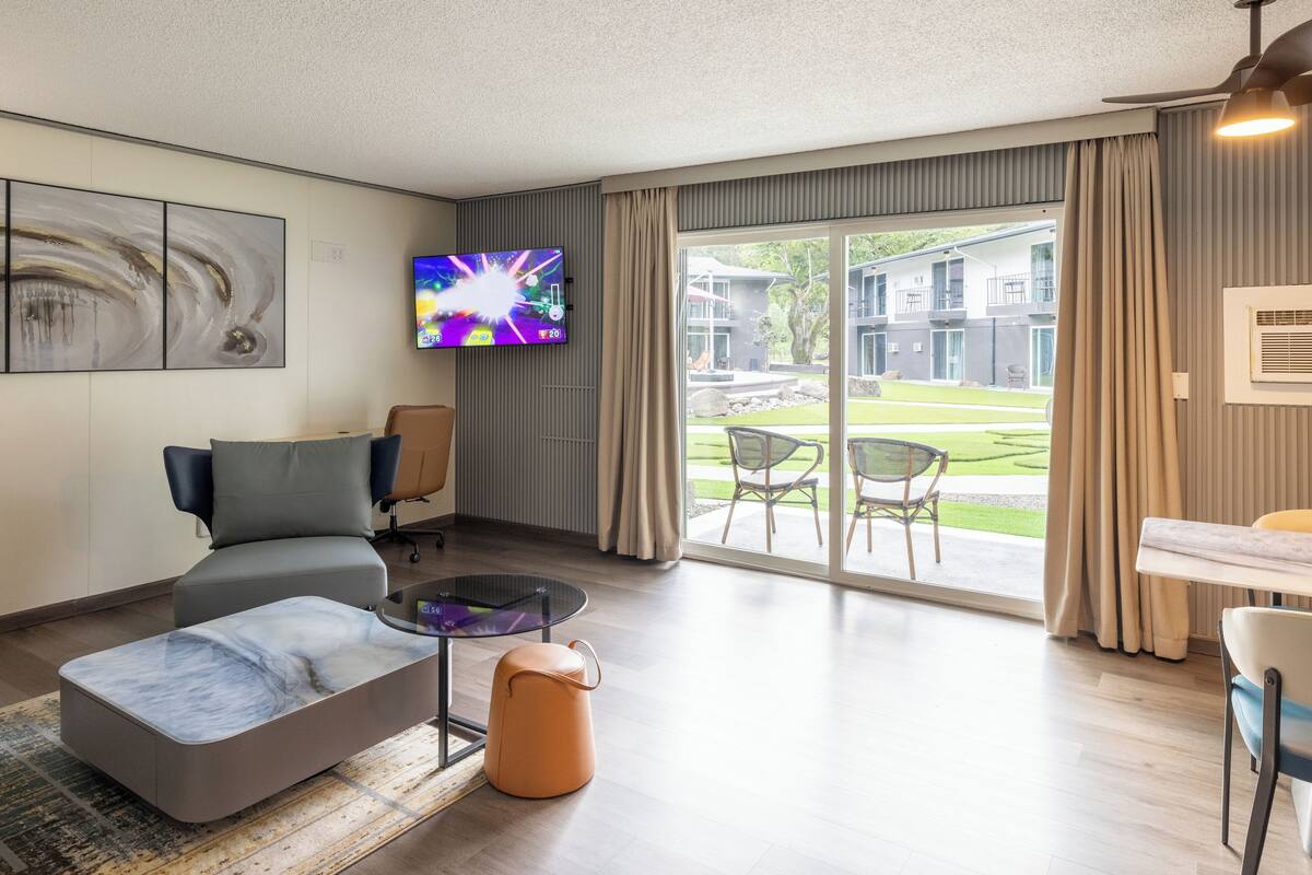 Una sala de estar moderna con un sofá, una mesa de centro y un televisor. Hay un ventanal corredero que da a un espacio exterior con césped y un edificio al fondo.