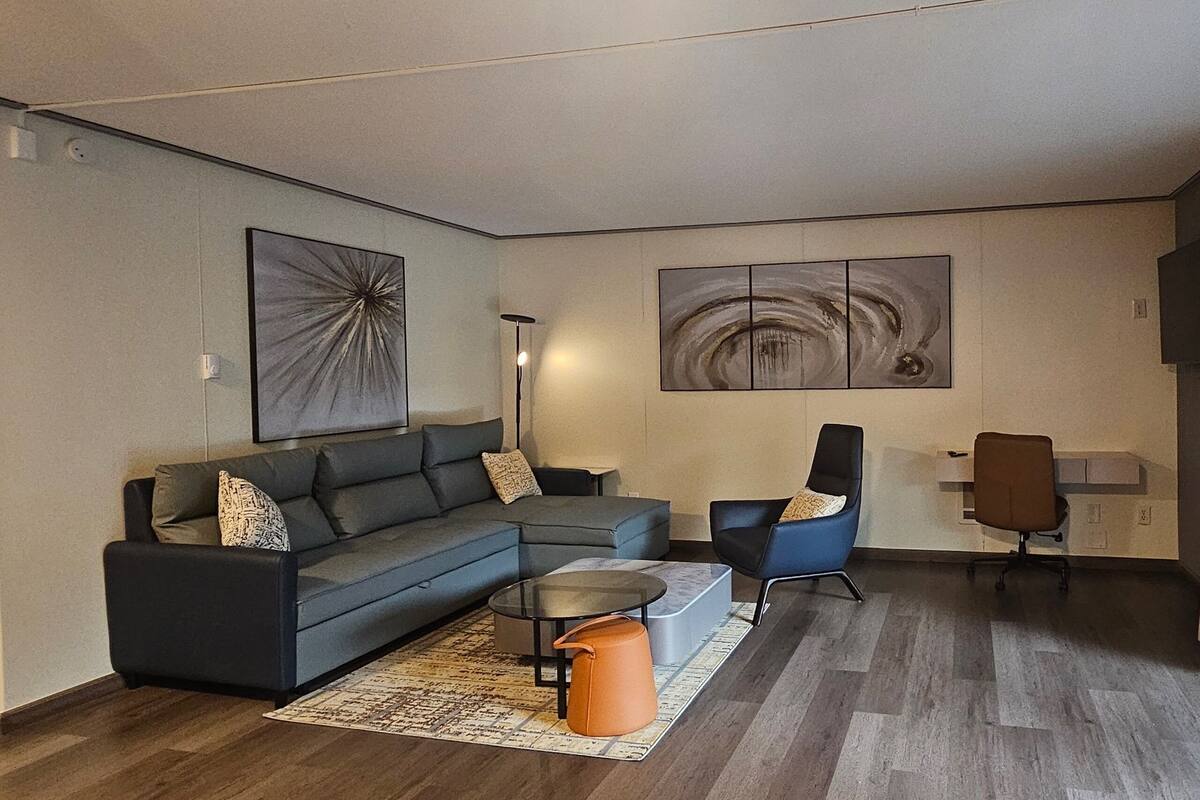 Una sala de estar moderna con un sofá seccional, una mesita de centro, un sillón azul y un amplio ventanal con cortinas.