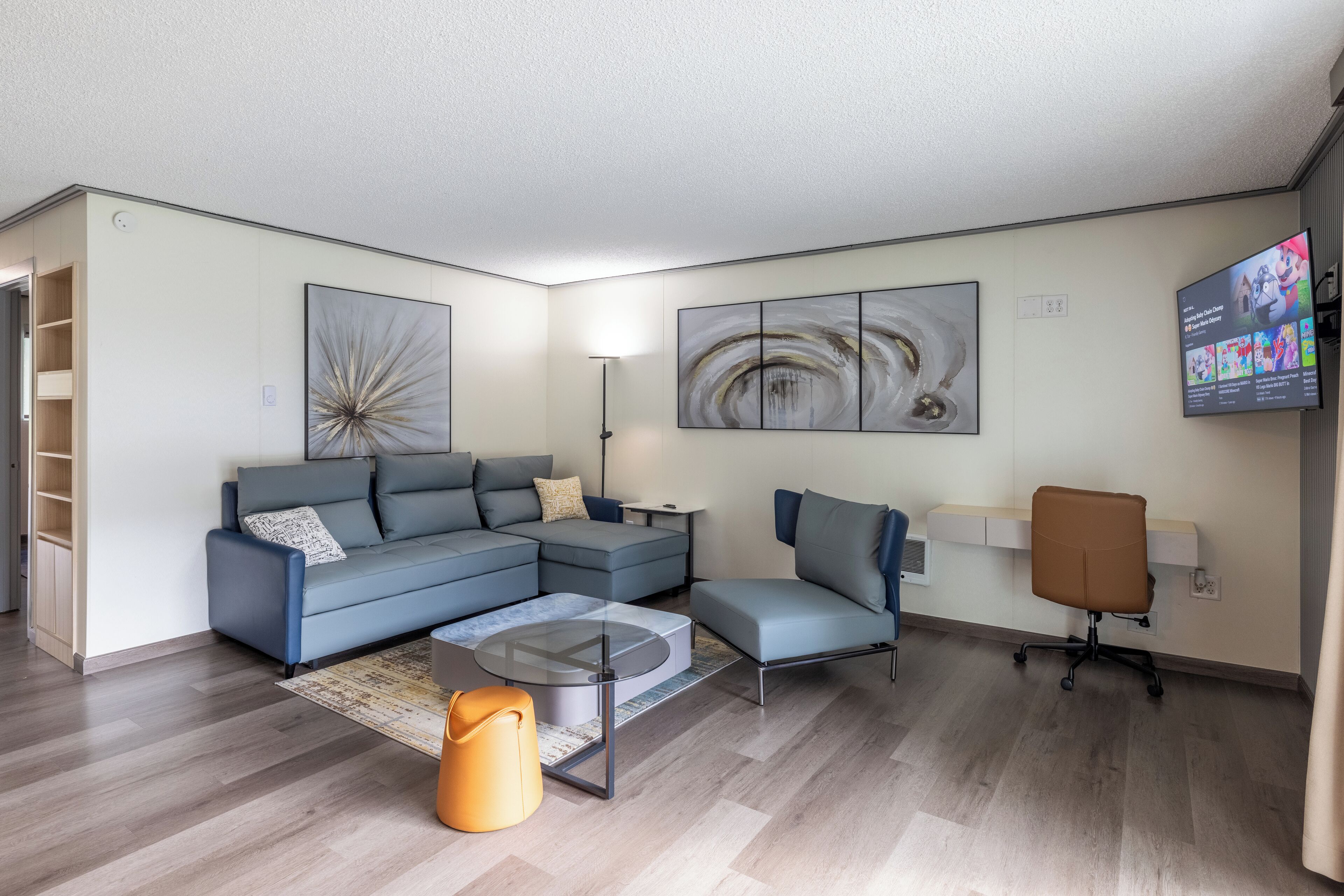 Un salon moderne avec un canapé d’angle, une table basse en verre, un fauteuil de bureau marron et un téléviseur à écran plat fixé au mur.
