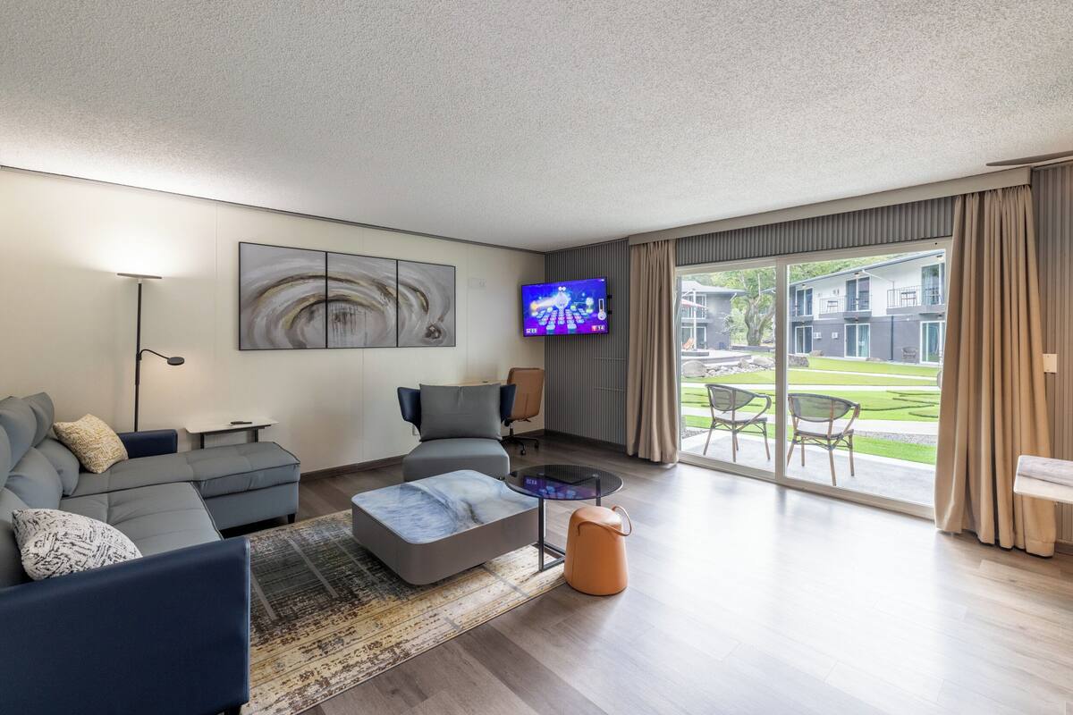 Una sala de estar moderna con un sofá modular, una mesa de centro y un televisor de pantalla plana montado en la pared. Se observa un ventanal con vistas a un edificio y un jardín.