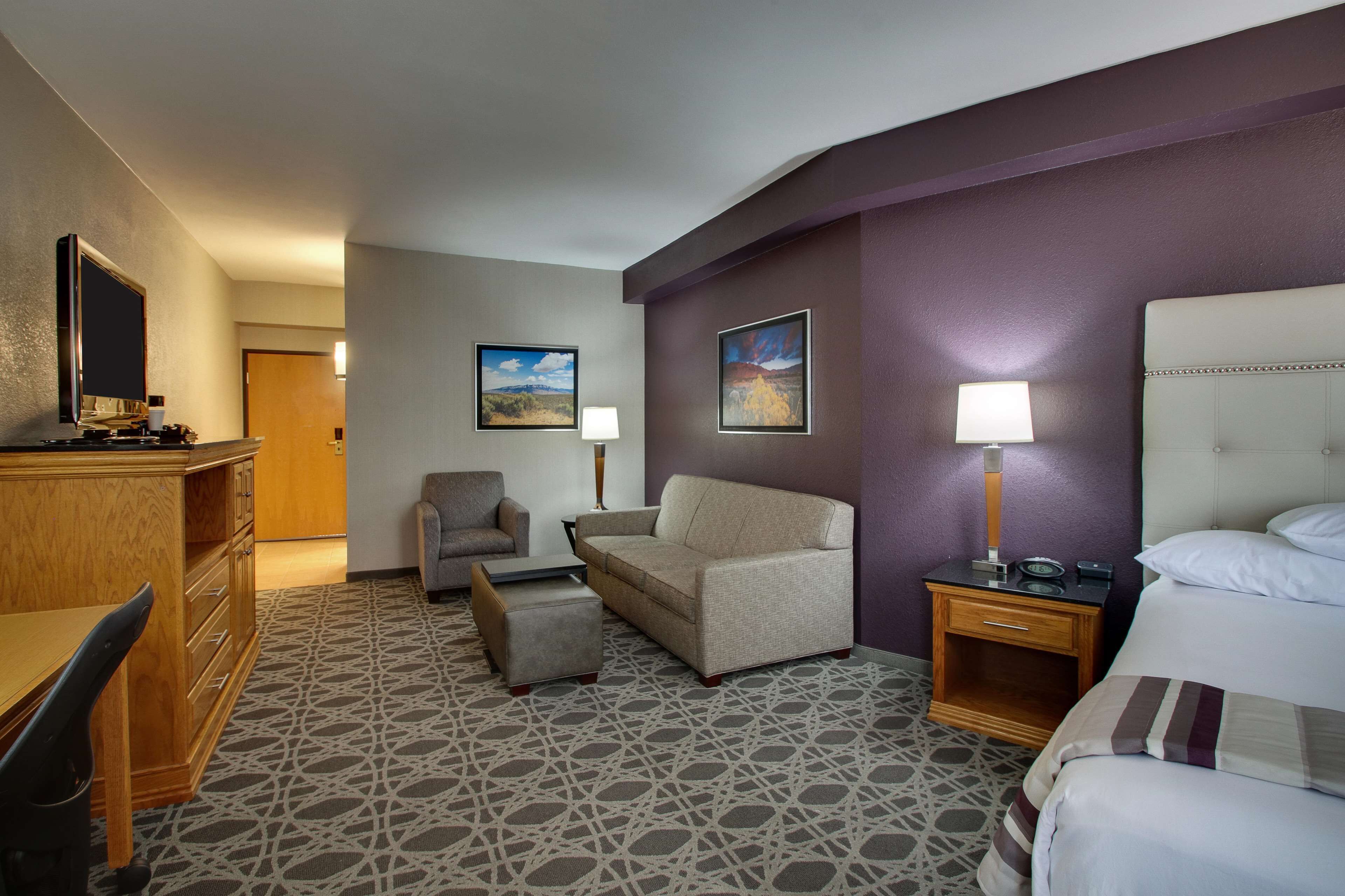 A hotel room with a bed, a desk, a chair, and a sofa. There are two paintings on the wall and a lamp on the nightstand.