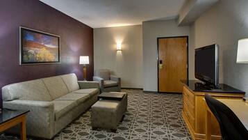 A hotel room with a sofa, armchair, ottoman, desk, television, and a painting on the wall.