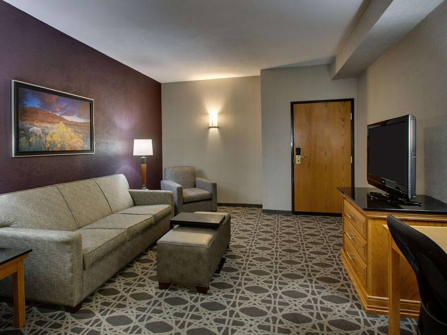 A hotel room with a sofa, armchair, ottoman, desk, television, and a painting on the wall.