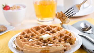 Un gaufre avec du beurre, servie sur une assiette, un verre de jus d’orange et un bol de yaourt garni d’une fraise.