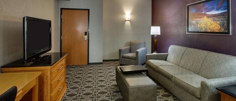 A hotel room with a flat-screen TV, a sofa, a chair, a small ottoman, and a wooden desk.