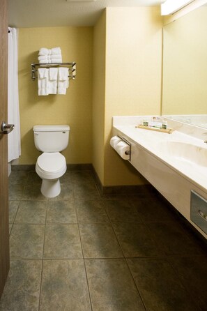 A bathroom with a toilet, a towel rack with folded towels, a sink with a mirror, and a toilet paper holder. - Merit Hotel & Suites (Fort McMurray)