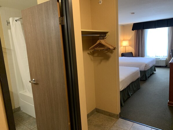 Habitación de hotel con puerta de madera, ducha, cama con cubrecolchón verde, mesita de noche y ventana con cortinas.