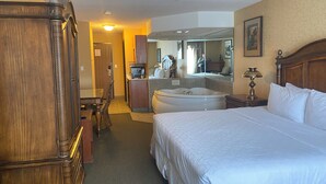 A hotel room with a large bed, a wooden wardrobe, a desk with a chair, a bathroom with a bathtub, and a mirror on the wall.