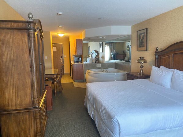 A hotel room with a large bed, a wooden wardrobe, a desk with a chair, a bathroom with a bathtub, and a mirror on the wall. - Merit Hotel & Suites (Fort McMurray)