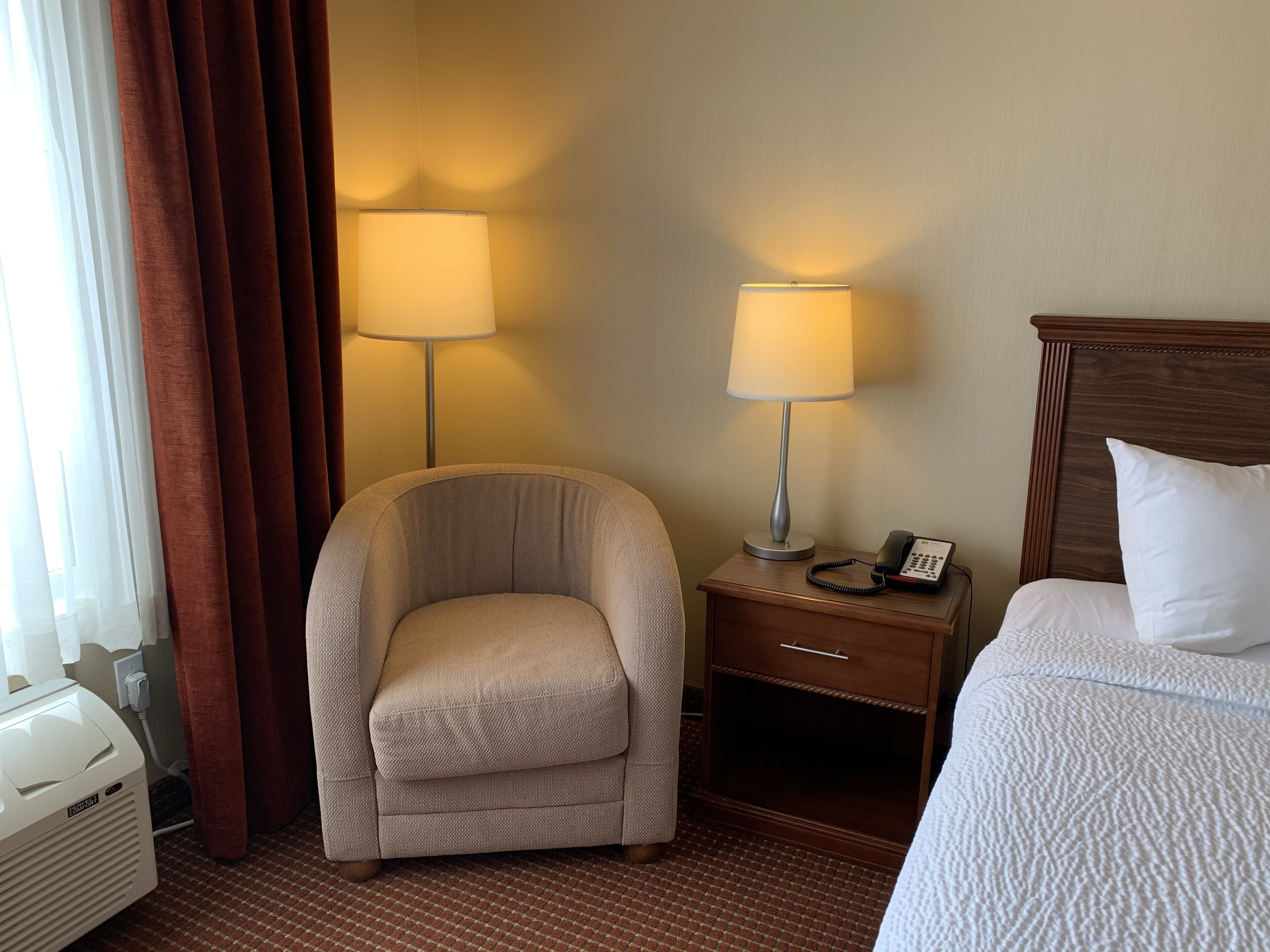 A hotel room with a bed, a chair, a bedside table with a lamp, and a power outlet.