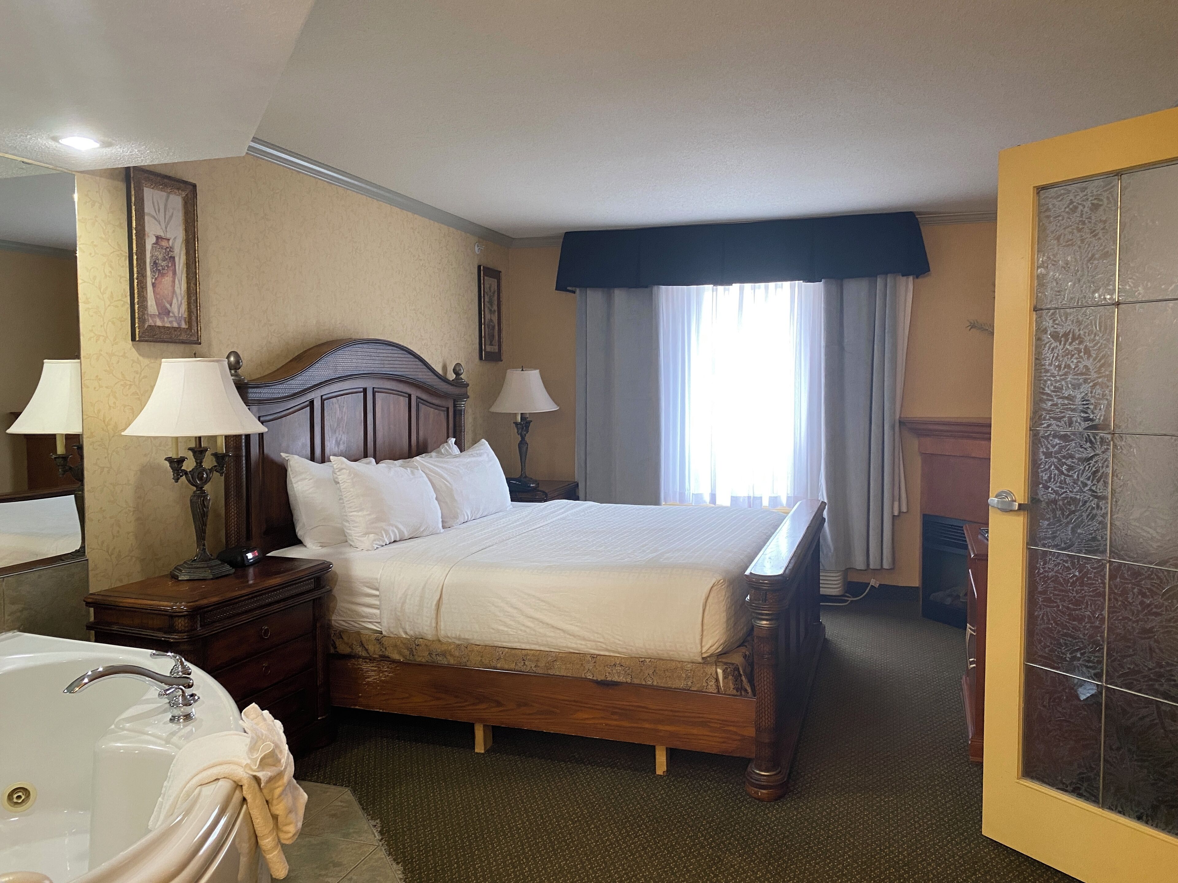 Quarto de hotel com cama de madeira, duas luminárias de cabeceira, mesa de cabeceira, banheiro com banheira e porta com painéis de vidro fosco.