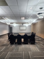 Una sala de conferencias con una larga mesa, sillas negras, piso alfombrado y una pantalla de proyector en la pared.