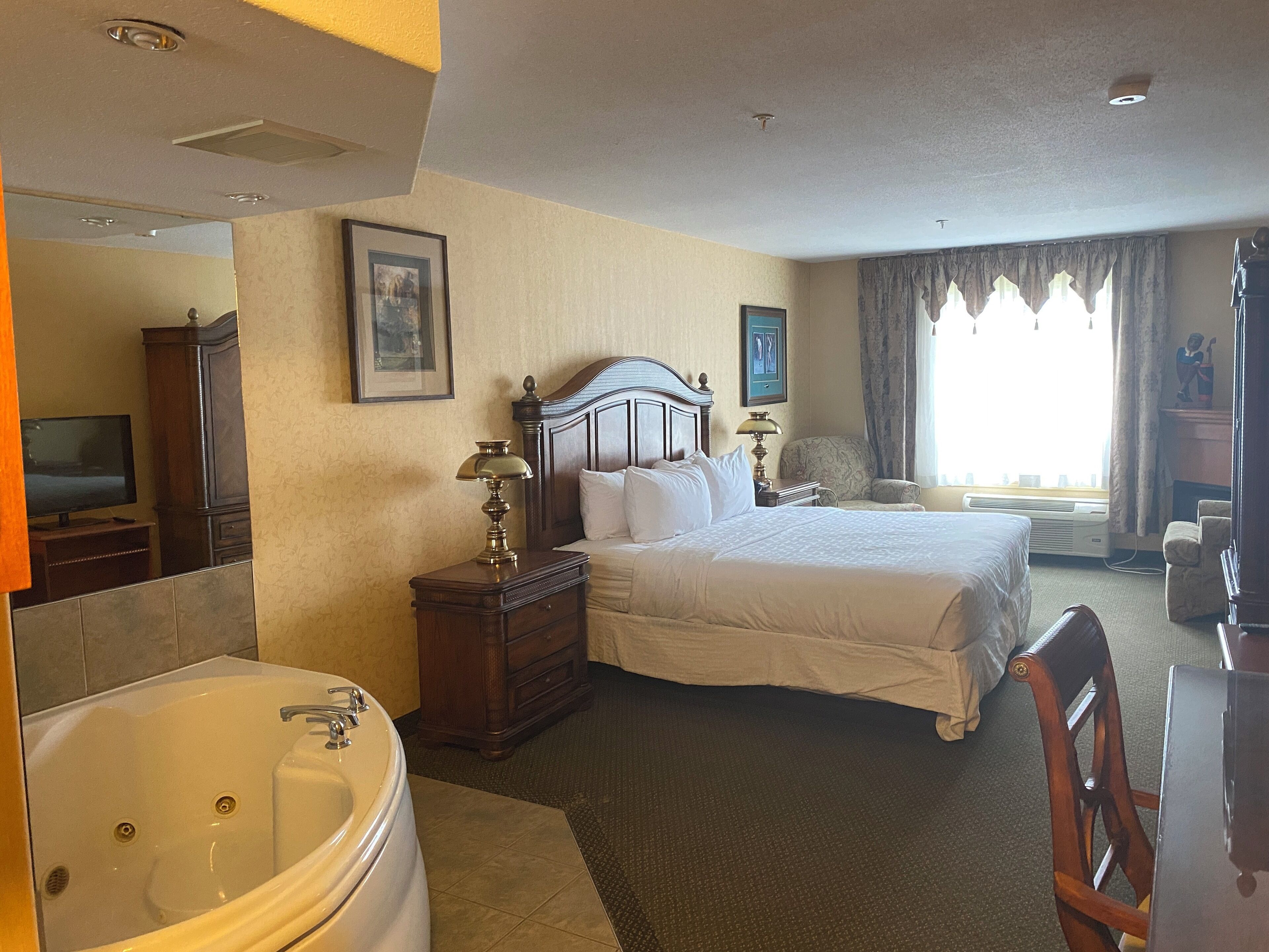 A hotel room with a large bed, a sitting area with a chair, a desk, a television, a cabinet, and a bathtub.