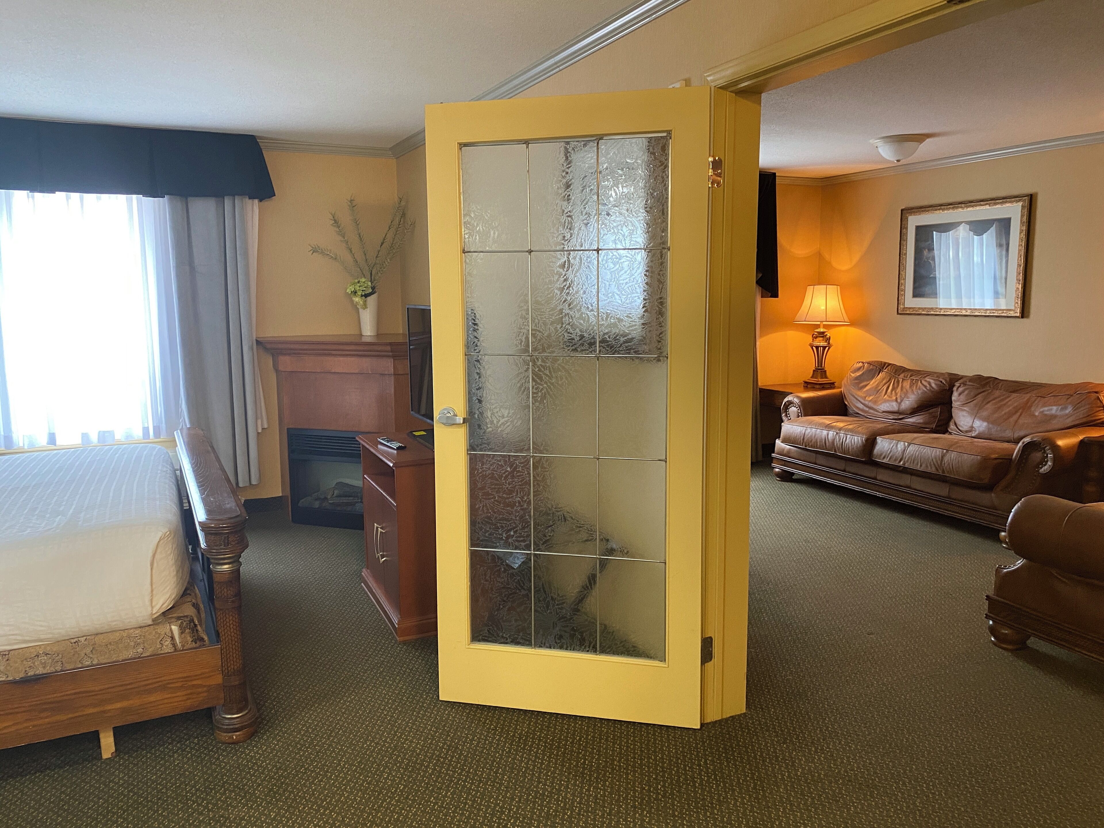 Una habitación de hotel con una cama, un aparador de madera, un sofá de cuero marrón y una puerta amarilla con paneles de vidrio esmerilado.