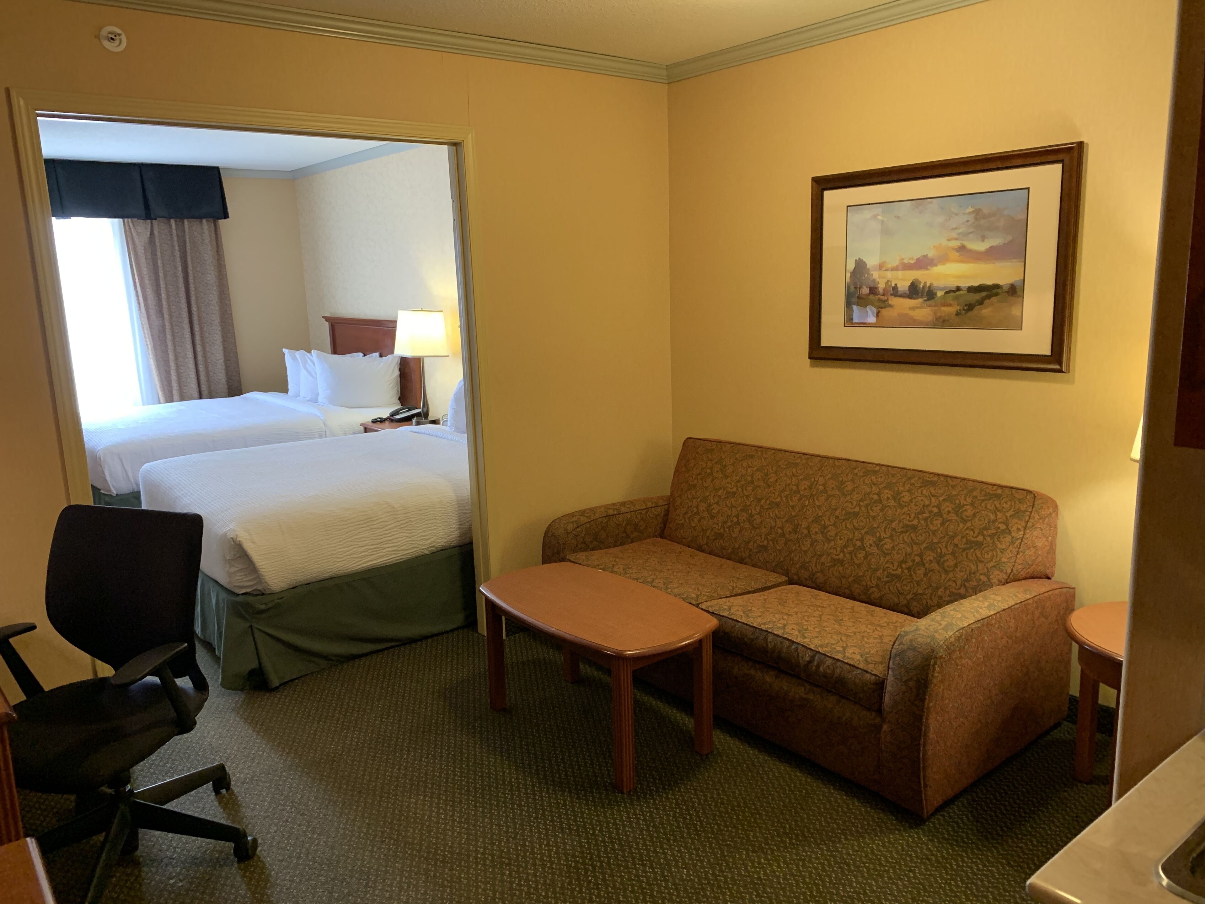 Una habitación de hotel con cama, sofá, una mesita, una silla y un cuadro en la pared.