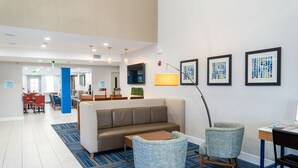 A modern hotel lobby with a sofa, chairs, a coffee table, a TV, framed artwork, and a desk. - Holiday Inn Express & Suites Lake Forest by IHG (Lake Forest)