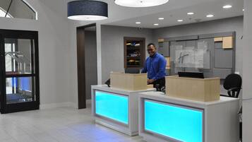 Eine Hotelrezeption im modernen Design, mit einem Empfangstresen mit digitaler Anzeige, einer Treppe und einer an der Wand befestigten Infotafel.