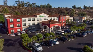 A multi-story hotel with a red and white facade, a central courtyard, and a parking lot with various vehicles. - Holiday Inn Express & Suites Lake Forest by IHG (Lake Forest)