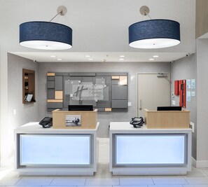 A modern hotel reception area with two reception desks, a large digital display, and a wall-mounted sign. - Holiday Inn Express & Suites Lake Forest by IHG (Lake Forest)